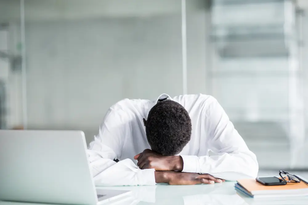 A person in a white shirt resting their head on their folded arms at a desk, with a laptop and documents nearby, illustrating the concept of 'Chronic Fatigue.'