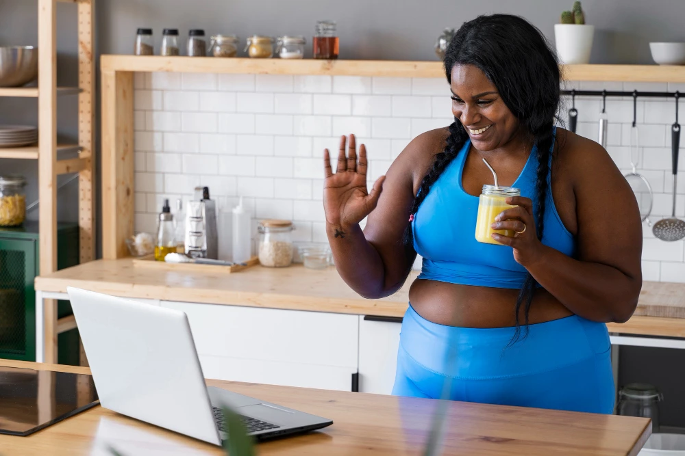 Happy woman in blue activewear enjoying a healthy smoothie while video chatting in a modern kitchen. Promoting weight loss membership in Katy, TX with a focus on wellness and healthy lifestyle support.