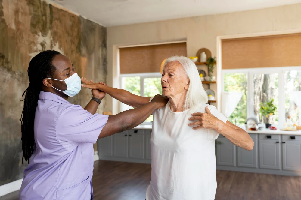 A healthcare professional wearing a lavender uniform and a face mask assists an elderly woman with physical therapy exercises in a well-lit home setting. The elderly woman, dressed in a white t-shirt, raises her arms as part of the therapy session while the caregiver provides support.