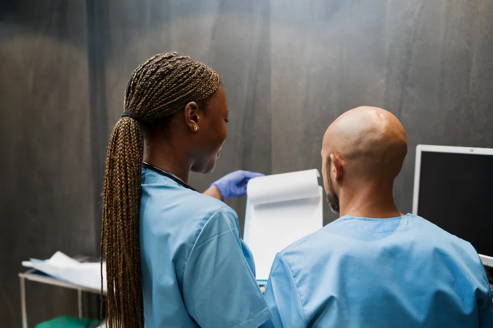 Two healthcare professionals, a male and a female, both dressed in blue medical scrubs, review a patient’s chart in a clinical setting with medical equipment in the background.