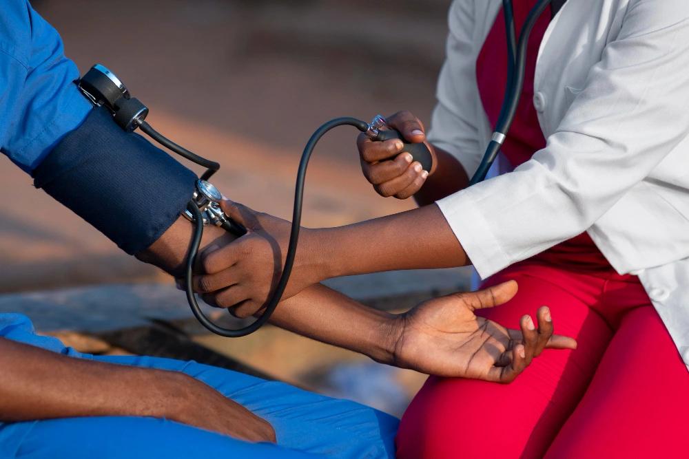 A healthcare professional wearing a white coat and red scrubs is checking a patient’s blood pressure using a manual sphygmomanometer. The patient is seated and wearing a blue outfit, extending their arm for the check-up. This represents chronic care, highlighting ongoing medical management for conditions like hypertension.