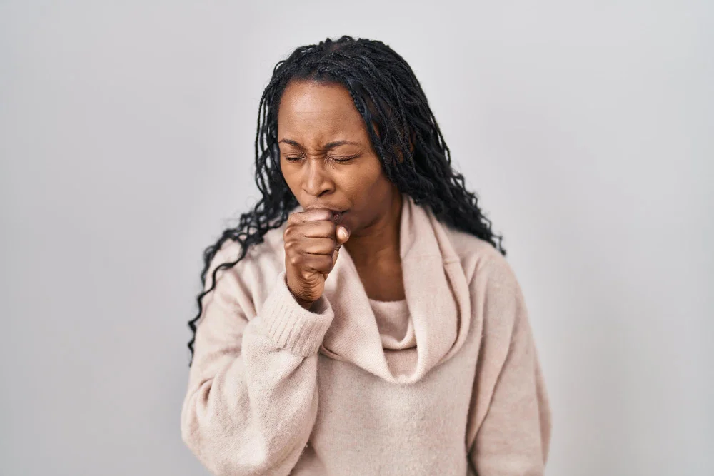 A middle-aged woman wearing a beige sweater coughs into her fist, appearing to be experiencing respiratory discomfort.