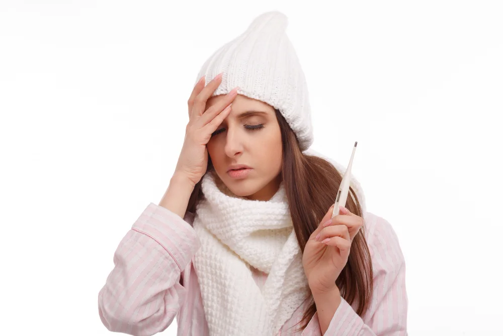 A woman dressed in winter clothing holds a thermometer while touching her forehead, displaying signs of fever and illness.