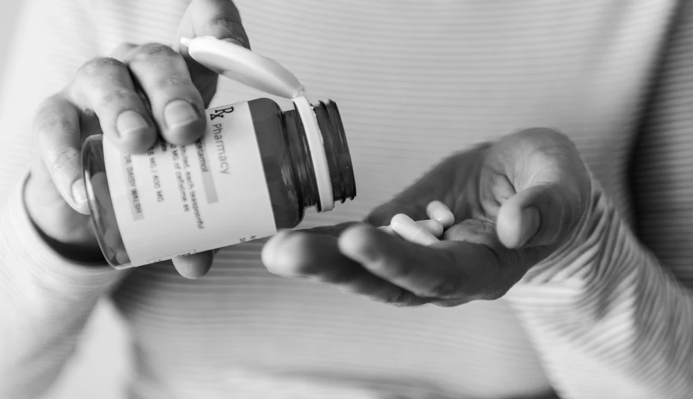 A close-up black and white photograph of a person pouring white pills from a prescription bottle labeled "Rx Pharmacy" into their palm, representing medication use and healthcare.