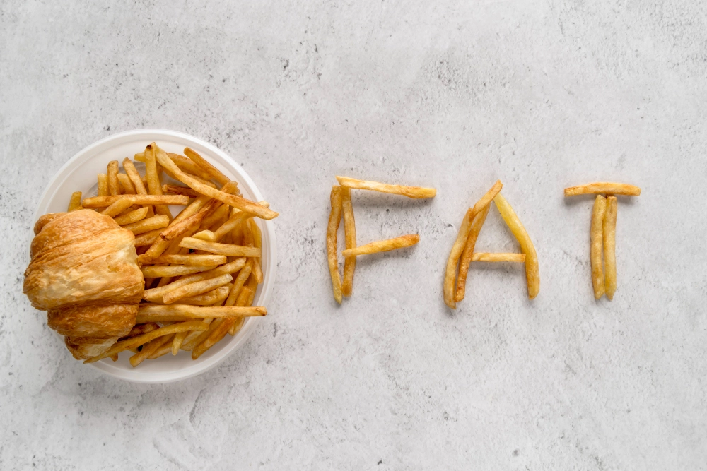 A plate of french fries and a croissant with the word "FAT" spelled out using fries on a gray surface.