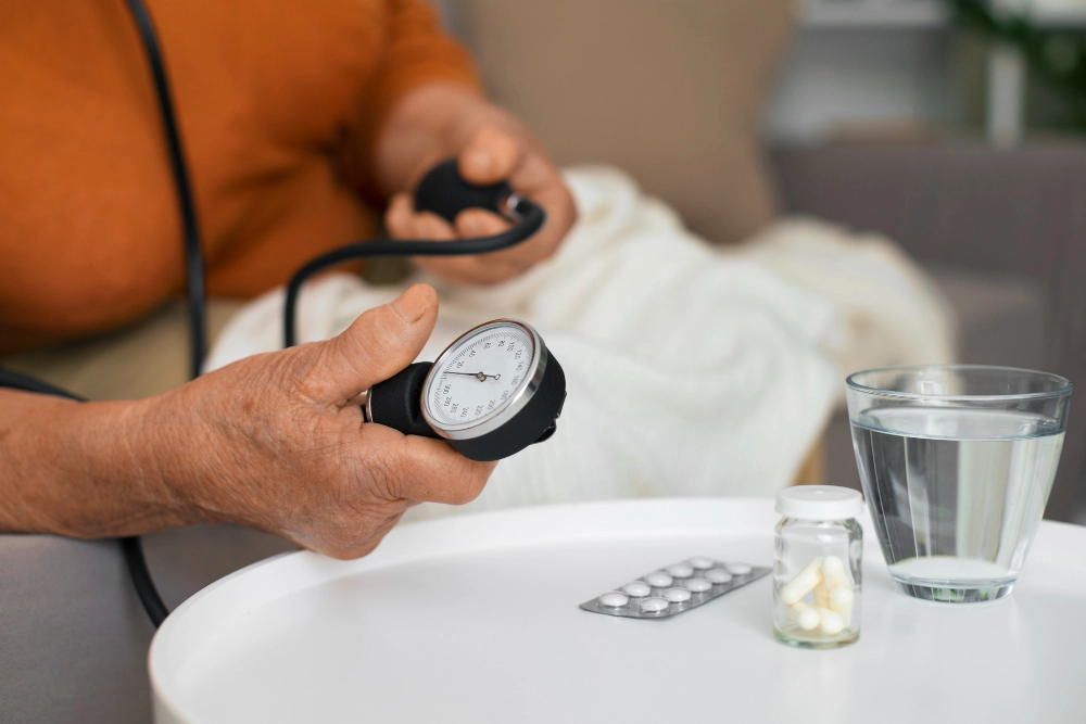 An elderly person holding a blood pressure monitor while sitting at a table with a glass of water and medication.