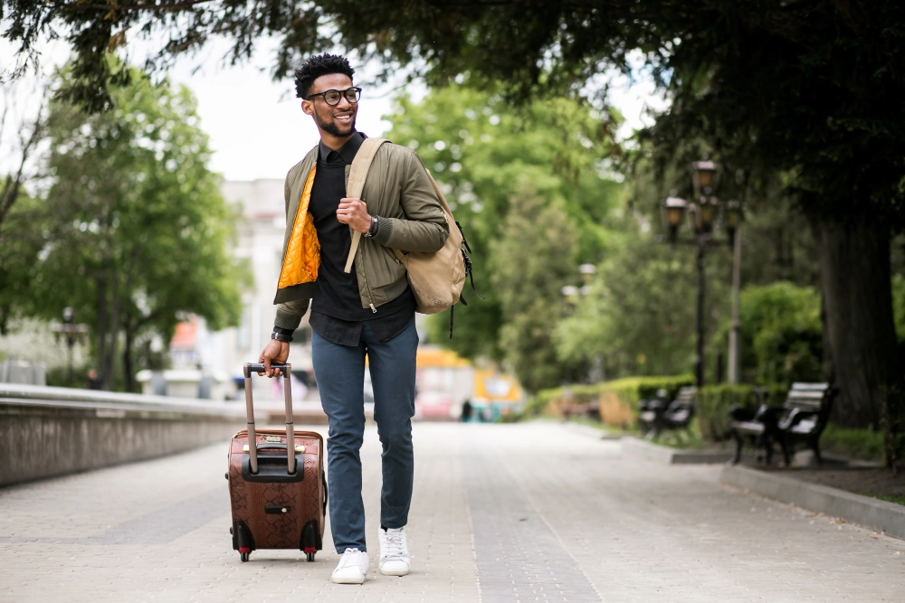 A young man with glasses, a backpack, and a rolling suitcase walks confidently along a tree-lined path, symbolizing travel and new beginnings, possibly related to immigration or relocation.