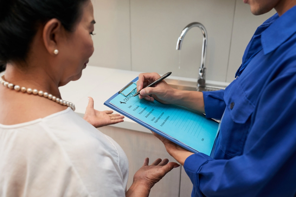 A close-up of a person signing an official document on a blue clipboard, possibly related to medical or immigration paperwork, with a woman in a white blouse and pearl necklace listening attentively.