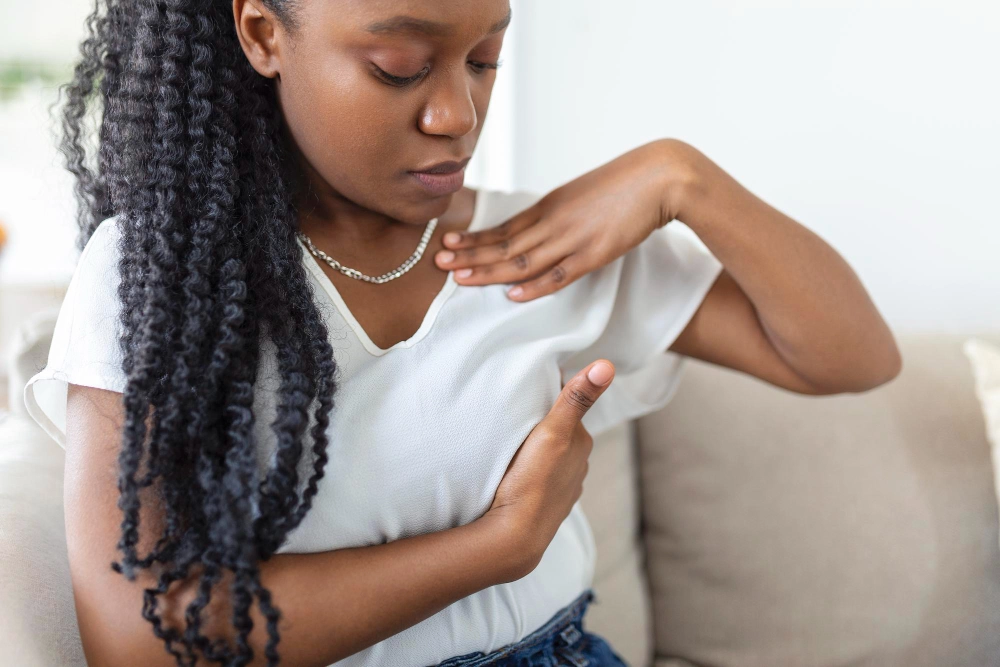 A young woman in a white top performing a breast self-exam by pressing her fingers against her chest while sitting on a couch.