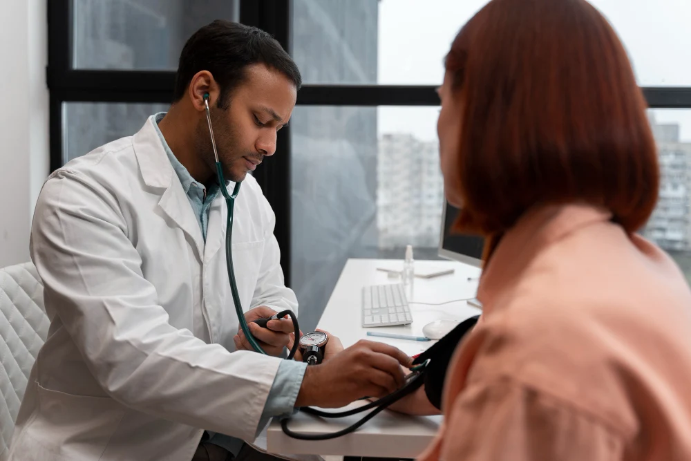 A doctor in a white coat using a stethoscope to check a female patient’s blood pressure in a modern medical office with a window view of a cityscape in the background.