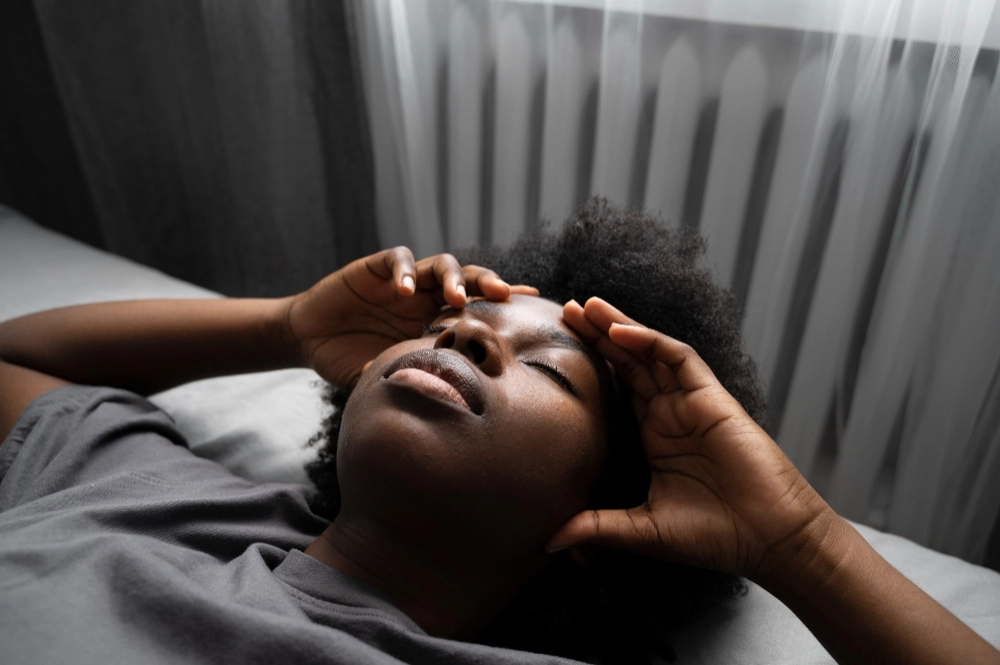 A woman lying in bed with her eyes closed, holding her head as if experiencing a headache or fatigue.