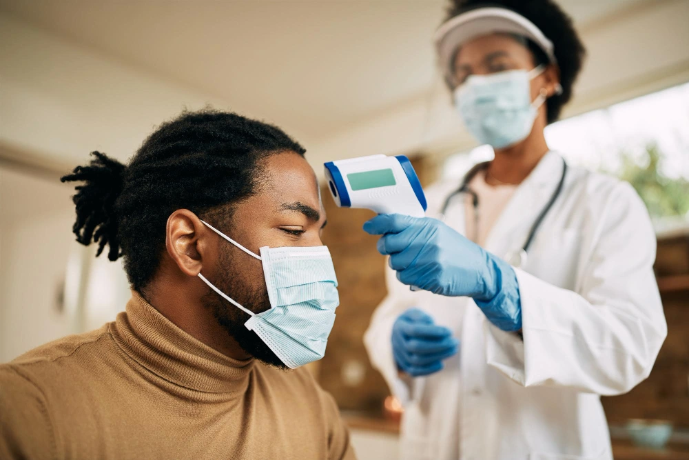 A healthcare professional wearing a face mask and gloves uses a contactless infrared thermometer to check the temperature of a patient, who is also masked. The scene illustrates preventive care measures in a medical setting.