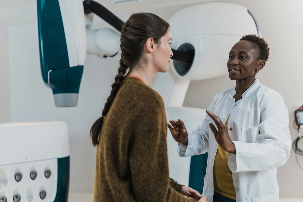 A female doctor wearing a white lab coat is explaining a medical procedure to a female patient with long braided hair in a clinical setting.