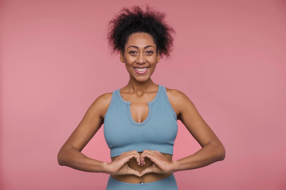 A smiling woman in athletic wear stands against a pink background, forming a heart shape with her hands over her abdomen. The image represents wellness, self-care, and women's health.