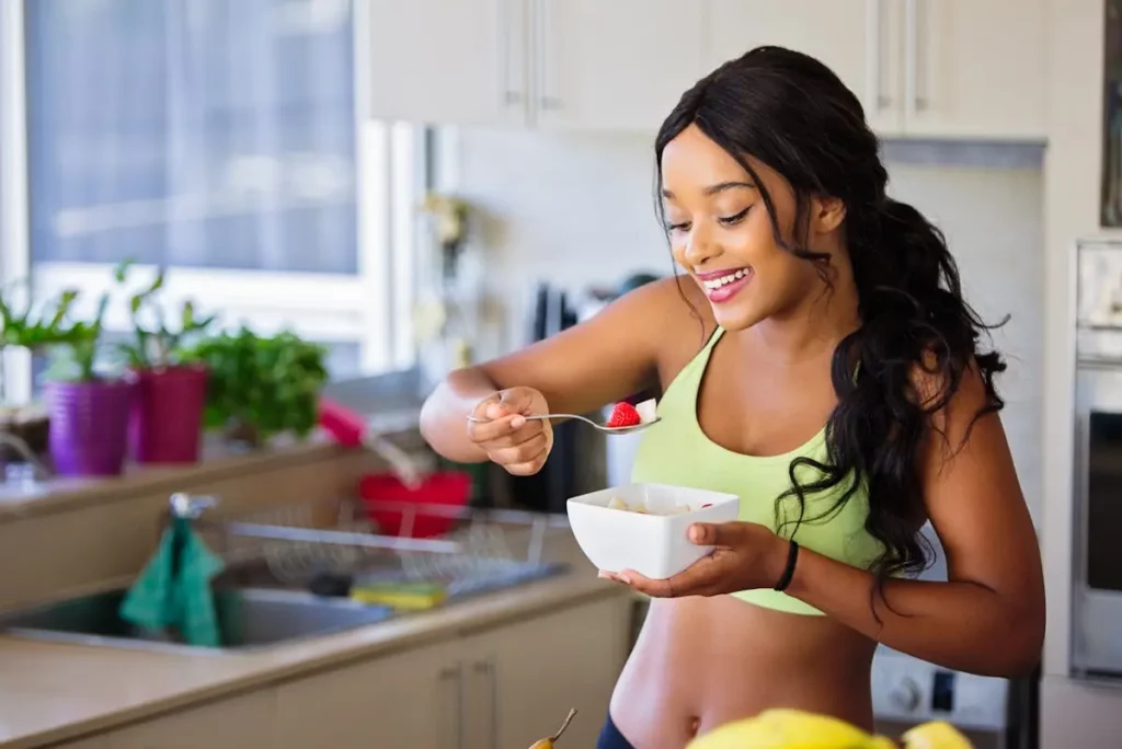 A smiling woman in a bright kitchen wearing a green sports bra, enjoying a healthy bowl of fresh fruit and yogurt, emphasizing mindful eating and wellness.