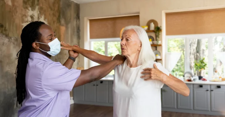 A healthcare professional wearing a lavender uniform and a face mask assists an elderly woman with physical therapy exercises in a well-lit home setting. The elderly woman, dressed in a white t-shirt, raises her arms as part of the therapy session while the caregiver provides support.