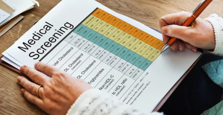 A person is holding a pen and reviewing a printed Medical Screening checklist. The document contains a table with different cholesterol and lipid levels, categorized into Optimal, Intermediate, and High ranges.