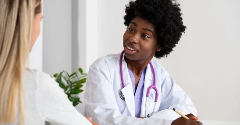 A female doctor wearing a white coat and a stethoscope around her neck attentively listens to a female patient during a medical consultation in a bright and modern clinic setting.