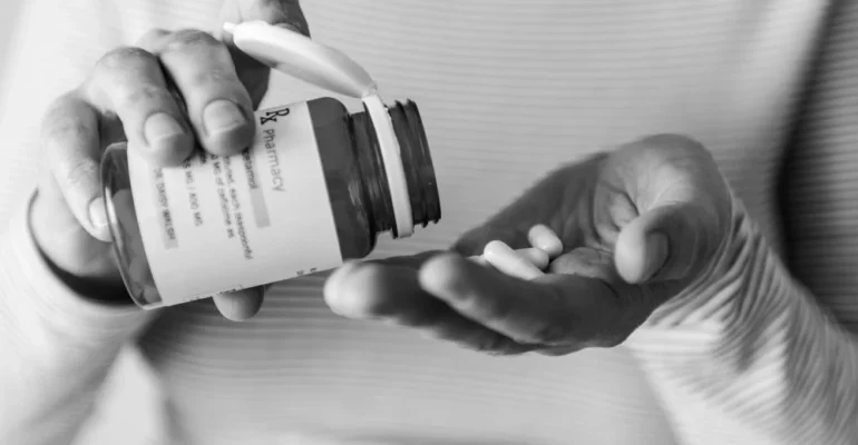 A close-up black and white photograph of a person pouring white pills from a prescription bottle labeled "Rx Pharmacy" into their palm, representing medication use and healthcare.