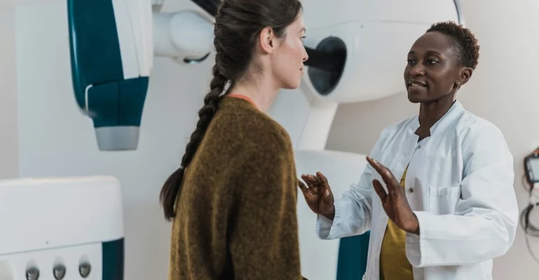 A female doctor wearing a white lab coat is explaining a medical procedure to a female patient with long braided hair in a clinical setting.