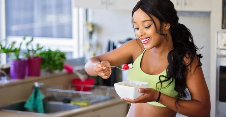 A smiling woman in a bright kitchen wearing a green sports bra, enjoying a healthy bowl of fresh fruit and yogurt, emphasizing mindful eating and wellness.